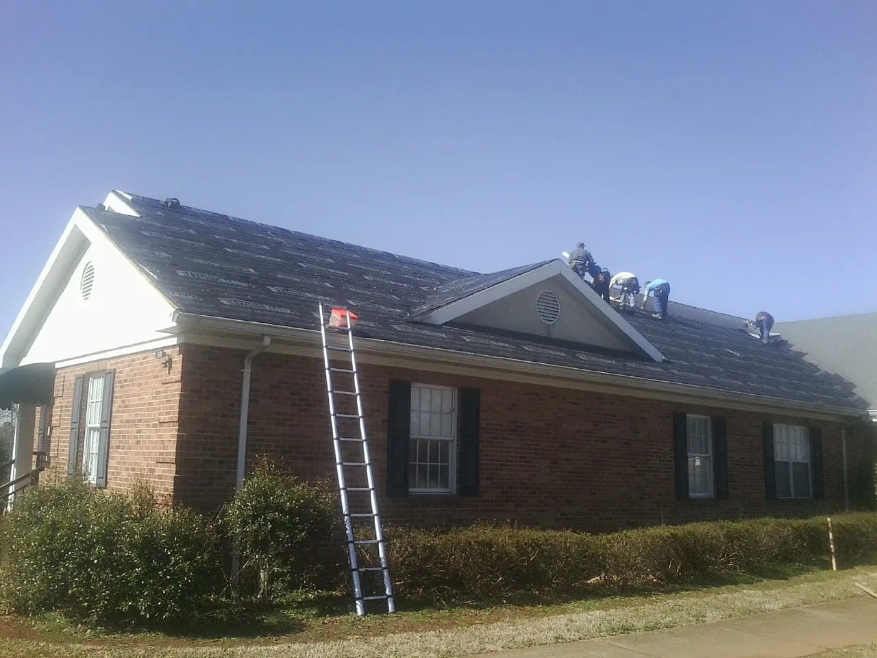Crew tearing shingles off a brick ranch roof with a ladder set — Metal Roof Repair work by our crew in Lyons
